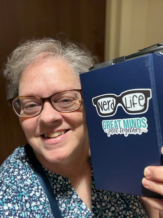 The author with a journal that has two stickers: "Nerd Life" and "Greaat Minds plot together."